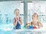 Et smilende barn og en voksen leger sammen i en pool under et forhæng af vandsprøjt, mens de holder farverige legetøjsbåde.
