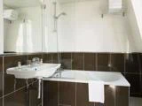 Et moderne badeværelse med badekar, bruseafskærmning i glas, væghængt håndvask, spejl og hylder med pænt foldede hvide håndklæder. Mørkebrune og hvide fliser pryder væggene.