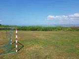 Et lille fodboldmål står på en græsmark ved siden af høje grønne siv, med en rolig blå sø og en klar himmel med spredte skyer i baggrunden.