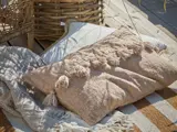Beige tekstureret pude med kvaster på et tæppe ved siden af en flettet kurv på en udendørs træterrasse.