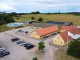Luftfoto af en landejendom med gule bygninger, parkerede biler, flagstænger og en swimmingpool, omgivet af marker og træer under en delvis overskyet himmel.