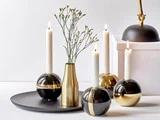 En moderne bordopstilling med tændte hvide lys i holdere i sort og guld, en guldvase med sarte blomster og en sort bakke på en hvid baggrund.