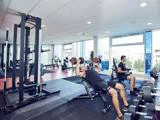 To mænd træner i et moderne fitnesscenter; den ene løfter håndvægte på en bænk, mens den anden bruger en stationær cykel. Vægtstativer og træningsudstyr er synlige i et lyst, rummeligt lokale.