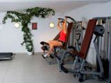 En kvinde i rød tanktop og shorts træner på en fitnessmaskine i et lyst rum med klinkegulv og en grøn plante i hjørnet.