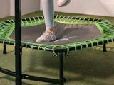 En person i lyse leggings og sneakers træder på en lille sort og grøn trampolin indendørs på kunstgræs.