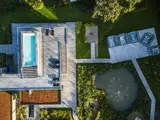 Luftfoto af et moderne hus med pool på taget, terrasse, grøn græsplæne, dam med springvand og udendørs siddepladser omgivet af træer og planter.