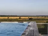 En udendørs pool med træterrasse har udsigt over marker og fjerne træer under en klar himmel.