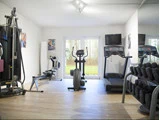 Et lyst træningscenter med træningsmaskiner, herunder løbebånd, crosstrainer, romaskine, vægtstativ og væghængt tv, der vender ud mod store glasdøre med udsigt til det fri.