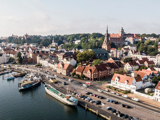 Luftfoto af en europæisk kystby med bygninger med røde tage, en kirke, parkerede biler og både, der ligger ved kajen.