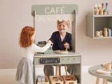 En ung dreng og en pige leger ved en legetøjscafé. Drengen sidder bag disken og smiler, mens pigen står foran og lader, som om hun vil købe noget. Legemad af træ står på disken.