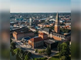 Luftfoto af et bybillede med røde murstensbygninger, et højt klokketårn og omkringliggende grønne områder under en klar himmel.
