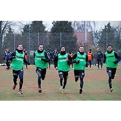 Fem fodboldspillere iført grønne træningstøj løber sammen på en græsbane under træning, med træer og andre spillere i baggrunden.