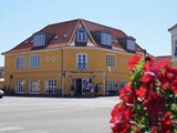 En gul toetagers hjørnebygning med kvistvinduer huser et hotel og en café. Røde blomster er slørede i forgrunden, og flere mennesker står nær indgangen.