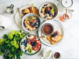 Et lyst morgenbord med skåle med havregryn toppet med frugt, vafler, en kop kaffe, mælk, honning og friske blomster på et hvidt bord.