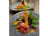 Et nærbillede af en gourmetret med farverige grøntsager, mikrogrønt og sauce, med en anden tallerken sløret i baggrunden.