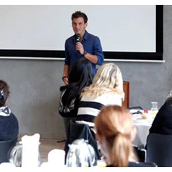 En mand står og taler i en mikrofon foran et siddende publikum på en konference eller et seminar med en projektionsskærm bag ham.