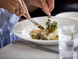 En person bruger kniv og gaffel til at skære i en anrettet gourmetret, pyntet med mikrogrønt, på et hvidt bord ved siden af et glas vand.