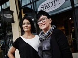 To kvinder står smilende uden for en tøjbutik med et skilt over sig, hvor der står SKOTT. Butikken har store vinduer og et moderne design.