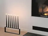 En moderne sort lysestage med seks tændte stearinlys står på et trægulv nær en pejs med en lille flamme indeni.