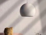 En moderne, kugleformet hvid pendel hænger fra loftet og kaster bløde skygger på en struktureret hvid væg. En lille sten og en plante er synlige nedenunder.