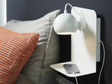 En moderne hvid væglampe med en indbygget hylde over en seng med en smartphone og et hvidt opladningskabel ved siden af en orange pude.