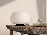 En rund, hvid lampe på et bord af ru sten ved siden af nogle bøger og en krystal, hvor det bløde lys kaster milde skygger på væggen.
