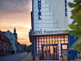 En hotelindgang med HOTEL Frederikshavn-skilt, blå indrammede glasdøre og planter på en stille gade ved solnedgang.