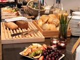 En morgenmadsbuffet med forskellige brød, rundstykker, krukker med korn, frisk frugt, tallerkener og stearinlys arrangeret på et bord.