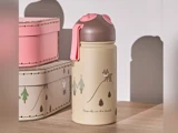 En beige vandflaske med lyserødt og brunt låg, dekoreret med træer og dyr, står på en træplade ved siden af to stablede lyserøde og beige kasser.