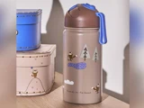 En beige vandflaske med brunt låg og blåt håndtag, dekoreret med søde vaskebjørne- og skovmotiver, står ved siden af blå og beige kasser.