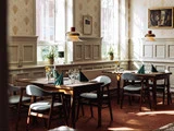 Solbeskinnet restaurant med pænt dækkede træborde, grønne servietter, blå stole, potteplanter i vindueskarmene og indrammet kunst på cremefarvede panelvægge.