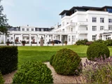 En stor hvid hotelbygning med balkoner og mange vinduer, omgivet af anlagte haver, grønne plæner, formklippede buske og blomstrende lavendler i forgrunden.