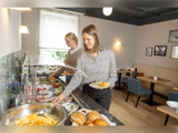 To kvinder serverer morgenmad for sig selv fra en buffet med røræg, kager, frugt og yoghurt i et lyst, moderne spiseområde.