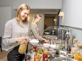 To kvinder serverer morgenmad for sig selv fra en buffet med morgenmadsprodukter, syltetøj, yoghurt og andre madvarer. Den ene kvinde hælder morgenmadsprodukter i en skål, mens den anden vælger mad fr