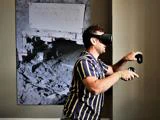 En mand i en stribet skjorte bruger et VR-headset og controllere, mens han står foran et stort sort-hvidt foto med håndskrevne noter.