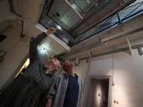 En kvinde og en mand står indendørs og kigger op og peger mod en netåbning i loftet, hvor der strømmer lys ned fra oven.