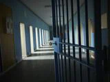 En svagt oplyst fængselsgang med en delvist åben metalport og sollys, der strømmer ind gennem sprossede vinduer langs den ene væg.