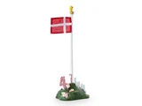 En hvid flagstang med et dansk flag, toppet af en lille gul and, står på en grøn base med blomster og gaver, omgivet af et hvidt stakit.
