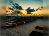 En tagterrasse af træ med oplyste trapper og bænke vises ved solnedgang med dramatiske skyer og orange og blå nuancer på himlen.