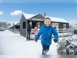 En smilende dreng i blå jakke løber mod kameraet foran sneklædte hytter, mens en familie leger sammen i sneen bag ham.