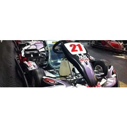 Nærbillede af en gokart med nummer 21 på fronten, parkeret indendørs på en bane med andre gokarts i baggrunden.