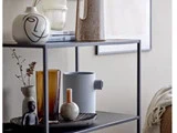 En moderne sort metalhylde med dekorative genstande, bl.a. abstrakte vaser, glas, en lille statue, et stearinlys og bøger arrangeret pænt på to hylder.