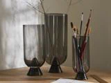 Tre røgfarvede glasvaser på en træplade; to indeholder tørrede grene, og en indeholder pensler. Blødt naturligt lys kaster skygger på væggen.
