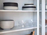 I et hvidt skab med glaslåge står der to hylder med stablede keramiske tallerkener, en stor skål og to skåle af klart glas.