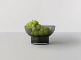 En skål af klart glas fyldt med grønne druer står på en hvid overflade foran en ensfarvet hvid baggrund.