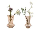 To lysebrune glasvaser med hver et par stilke af sarte hvide og lilla blomster med grønt løv står side om side på en hvid baggrund.