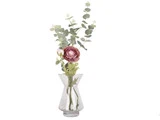 En klar glasvase rummer en pink ranunkelblomst med grønne blade og eukalyptusstængler på en hvid baggrund.