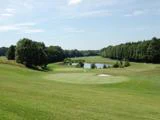 En grøn golfbane med en sandbunker, omgivet af træer og en dam i det fjerne under en delvist overskyet himmel.