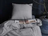 En seng med grå lagner og pude; et åbent magasin og briller ligger på betrækket, og der står en stor potteplante ved siden af sengen.