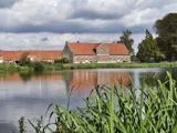 En stor bygning med et rødt tag står ved en rolig dam, omgivet af grønt græs og træer under en overskyet himmel.